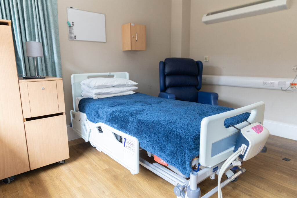 Heart of Kent Hospice Inpatient unit bedroom. There is a bed and blue armchair, bright windows with a view to the Hospice garden.